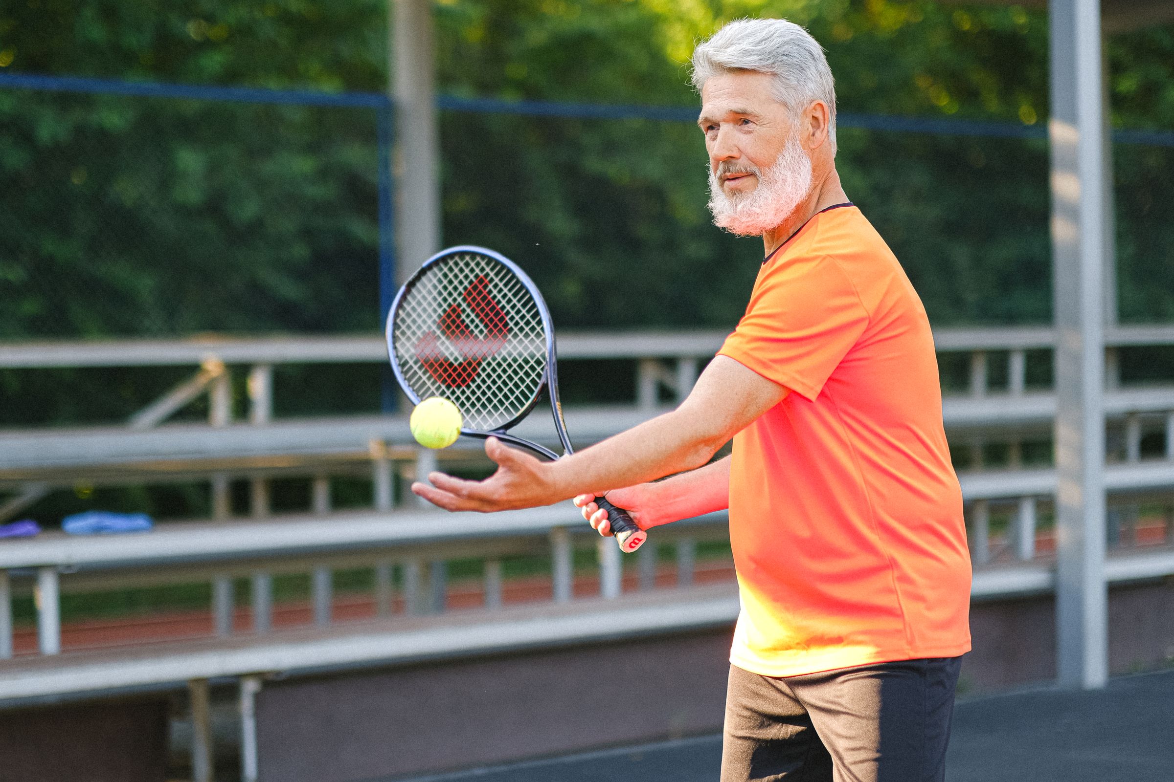 An older tennis player in an orange shirt preparing to serve in the sunshine